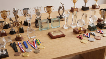 Vielfältige Sammlung verschiedener Pokale, Glas-Awards und Medaillen auf einer hellen Holzfläche – KI-generiertes Symbolbild