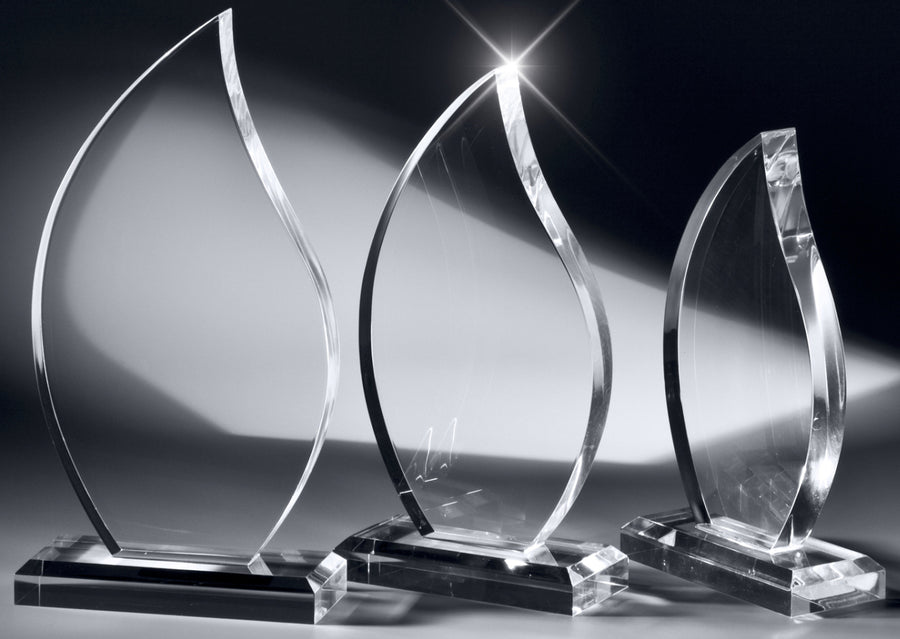 Drei klare, längliche Kristallpreise mit geschwungenen, blattähnlichen Formen auf einer reflektierenden Oberfläche vor einem dunklen Hintergrund, die die Präzision der Awards Osnabrück 3-er Serie 200 x 100 mm – 250 x 125 mm PK733603-01-3 zur Schau stellen.