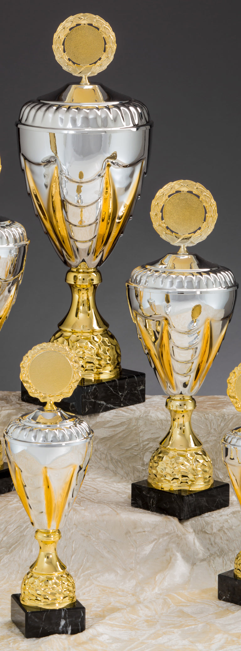 Mehrere silberne und goldene Trophäen in verschiedenen Größen mit runden Plaketten oben drauf, gefertigt aus hochwertigen Materialien, sind auf einer beige drapierten Oberfläche angeordnet und zeigen die Pokale Lemgo 3-er Pokalserie 275 mm - 346 mm