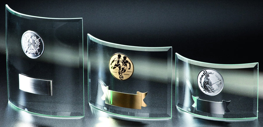 Drei klare Glasplaketten mit abgerundeten Kanten, jede mit einem anderen Metallemblem und einem leeren Gravurschild an der Unterseite, vor dunklem Hintergrund - Glaspokal Bochum 3-er Serie 100x145 mm - 174x162 mm PK766483-81-3-E50.