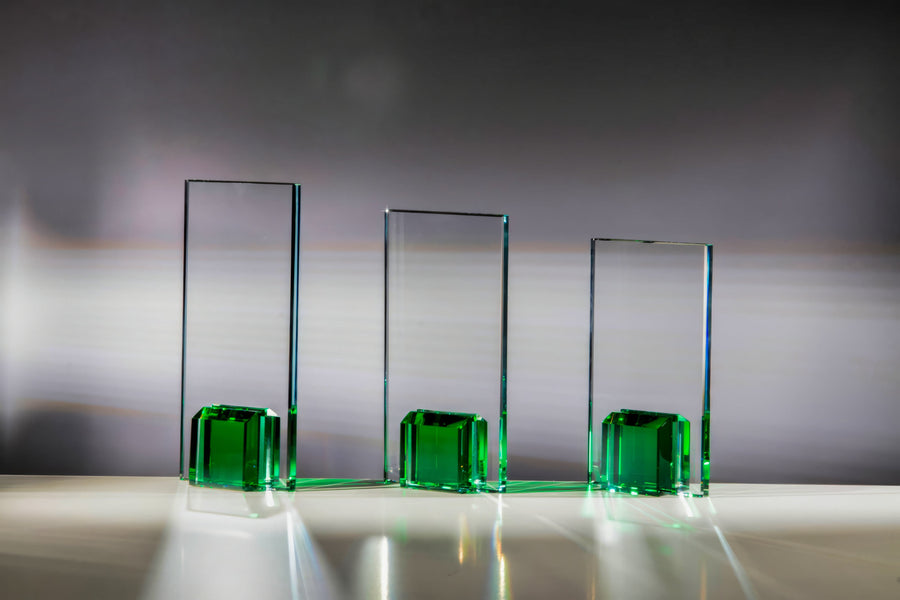 Drei rechteckige Glasprismen mit grüner Basis sind in einer Reihe auf einer reflektierenden Oberfläche mit Farbverlaufshintergrund platziert. Licht fällt durch sie hindurch und wirft grüne Reflexionen und Schatten, ähnlich wie Awards Hannover 3-er Serie 180x100 mm - 220x100 mm PK768134-32-3 komplizierte Lasergravurdetails im Scheinwerferlicht zur Geltung bringt.