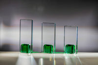 Thumbnail for Drei vertikale Glaspokal Bremen 3er Serie 180x100 mm – 220x100 mm PK768134-32-3-E50 mit grüner Basis sind auf einer weißen Oberfläche platziert, werfen Schatten und reflektieren Licht vor einem grauen Verlaufshintergrund und ähneln so der Eleganz eines Emblems.