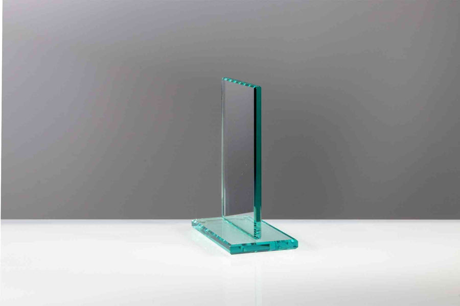 Ein transparenter Glaspokal Köln 4-er Serie 117x100 mm – 170x140 mm PK735265-60-4-E50 steht wie ein Emblem auf einer reflektierenden weißen Oberfläche vor einem schlichten grauen Hintergrund., glaspokale
