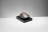 Thumbnail for Eine kleine, futuristische Skulptur mit geometrischen Formen, überwiegend in Grau mit bronzenen Akzenten, präsentiert auf einem rechteckigen schwarzen Sockel – eine perfekte Trophäe Computer Maus 70 mm x 150 mm