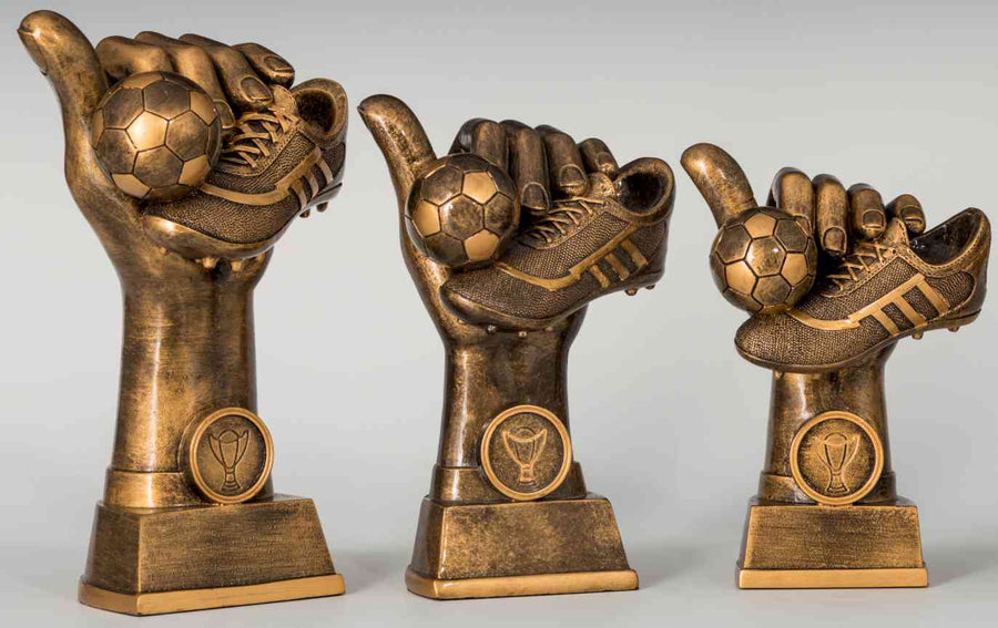 Drei bronzene Trophäen der 3-er Serie Fußball 130 mm - 172 mm PK739400-3 zeigen eine Hand, die einen Fußball und einen Stollen hält, jeweils auf einem rechteckigen Sockel mit einem runden Emblem an der Vorderseite des Sockels. Die aus hochwertigem Material gefertigten Trophäen variieren leicht in der Größe und zeigen ein exklusives Design.